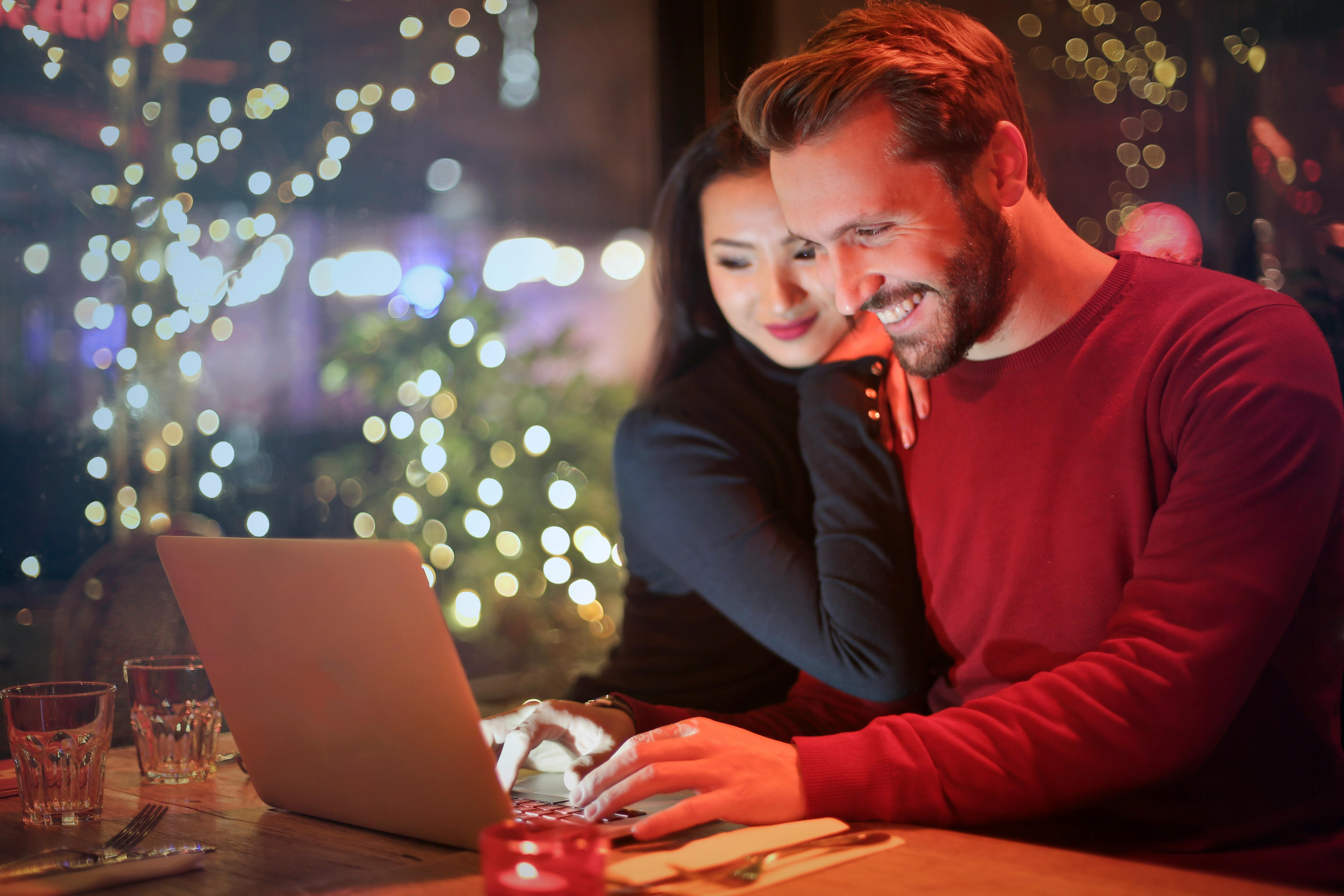 A man and woman are smiling, looking at a laptop. She is holding onto his shoulders.